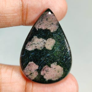 May include: A teardrop-shaped gemstone with a polished surface. The stone features a dark green base with patches of pink and white, creating a unique pattern. The gemstone is held between two fingers.