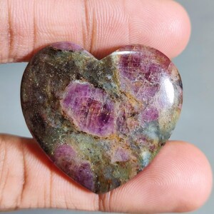 May include: A heart-shaped gemstone with a polished surface. The stone displays a mix of colours, including deep pink, brown, and green. The pink areas appear crystalline, while other areas have a mottled appearance. The gemstone is held in a hand.