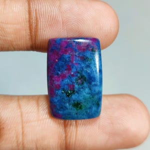 May include: A rectangular gemstone with a polished surface. The stone features a vibrant mix of colors, including deep blue, teal, magenta, and green. The colors are swirled and speckled throughout the stone, creating a unique pattern. The gemstone is held between two fingers.