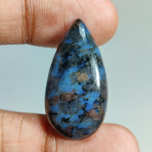 May include: A teardrop-shaped gemstone with a polished surface. The stone features a mix of blue, black, and brown hues, creating a marbled effect. The gemstone is held between two fingers, showcasing its size and shape.
