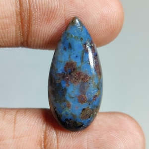 May include: A polished, teardrop-shaped gemstone. The stone has a striking blue base colour, with brown and dark blue inclusions. The gemstone is held between two fingers, highlighting its form and size.