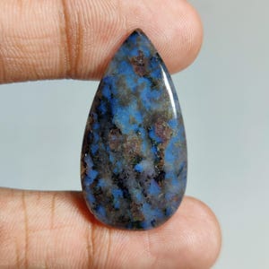 May include: A polished, teardrop-shaped gemstone. The stone displays a mottled pattern of blue, brown, and grey tones. The gemstone is held between two fingers, highlighting its size and form. The stone's colours and pattern create a unique, natural appearance. The stone is approximately 3 cm in length.