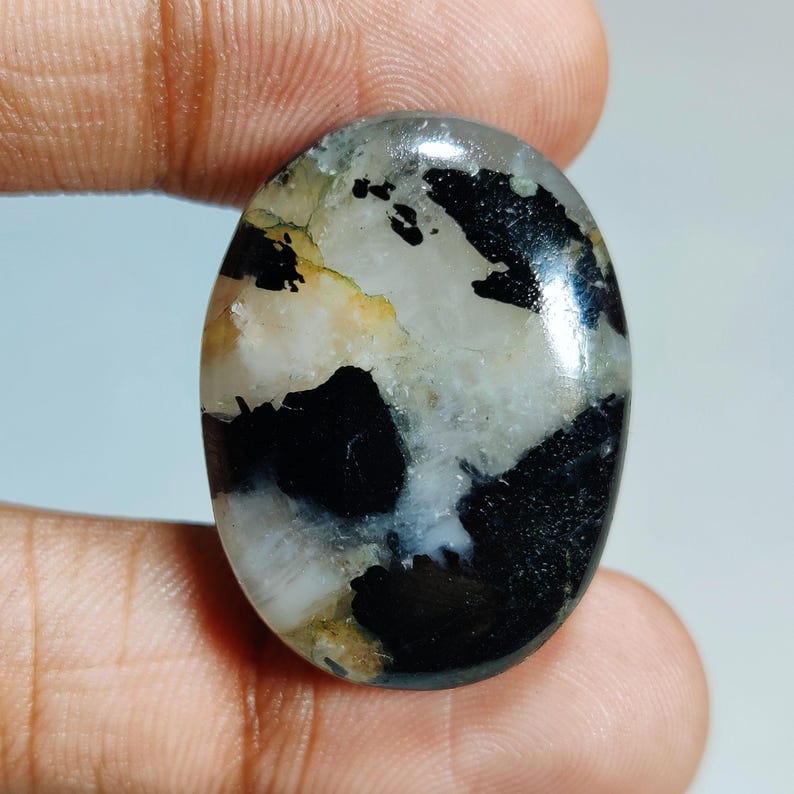 May include: Oval gemstone with a polished surface. The stone displays a mix of black, white, and grey colours, with some yellow and brown areas. The gemstone is held between two fingers.