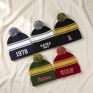 May include: Five knit beanies with pom-poms. One navy with white stripes and 1978, one black with white stripes and GAME DAY, one black with gold stripes and a pink ribbon, one olive with gold stripes and Pinktober, and one maroon with gold stripes.
