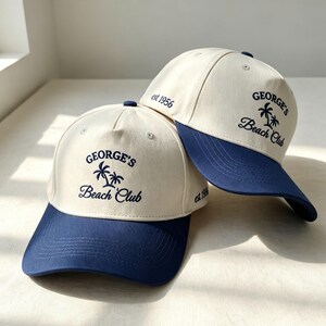 Puede incluir: Dos gorras de b&eacute;isbol con corona color crema y visera azul marino. Cada gorra presenta un bordado azul oscuro en la parte frontal que dice George's Beach Club con dos palmeras, y el texto est 1956 bordado en el panel lateral de las gorras.