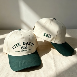 Puede incluir: Dos gorras de béisbol color blanco hueso con viseras verde oscuro. La gorra frontal presenta texto bordado en verde oscuro que dice THE BACH Club EST.2026. El lateral de la segunda gorra muestra el nombre Kait bordado en letra cursiva.