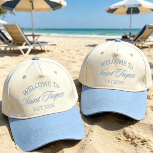 May include: Two baseball caps with a cream-colored crown and a light blue brim are displayed on a sandy beach. The caps have the embroidered text "WELCOME TO Saint Tropez EST.2026" in a matching light blue color.