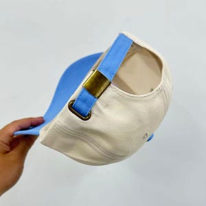 Op de afbeelding: Een baseballpet met een cr&egrave;mekleurige kroon en een lichtblauwe klep. De achterkant van de pet is voorzien van een lichtblauwe stoffen band met een verstelbare metalen gesp in antiek messing voor een goede pasvorm en een gestructureerd ontwerp.