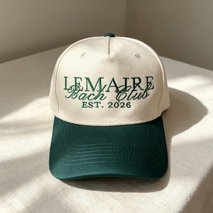 Pode incluir: Um bon&eacute; de beisebol cor de creme com aba verde-escura e bordado verde-escuro na frente. O texto diz LEMAIRE em letras de forma, Bach Club em letra cursiva e EST. 2026 abaixo. O bon&eacute; apresenta uma copa estruturada e um bot&atilde;o no topo.