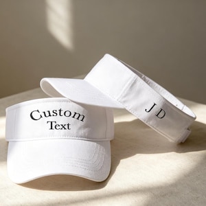 May include: Two white sun visors featuring black embroidered text. The front visor displays the words Custom Text in a serif font, while the second visor positioned behind it features the embroidered initials JD on the side panel.