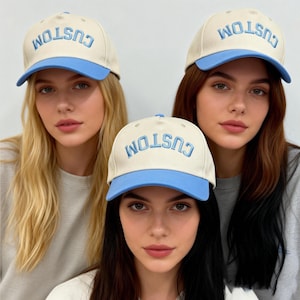 May include: Three baseball caps featuring a cream-colored crown and a light blue brim. Each cap displays the word CUSTOM embroidered in light blue, arched lettering across the front panel. The caps have a structured design with a button on top.