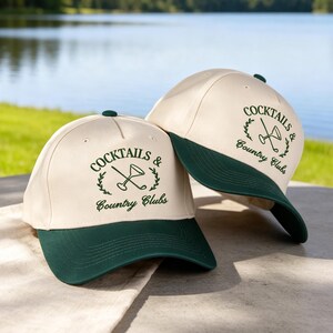 Puede incluir: Dos gorras de b&eacute;isbol de color crema con viseras verde oscuro y bordado verde a juego. La parte frontal de cada gorra presenta un gr&aacute;fico de una copa de martini cruzada con un palo de golf, rodeado de hojas de laurel y el texto Cocktails & Country Clubs.