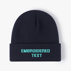 Puede incluir: Un gorro azul marino con las palabras "EMBROIDERED TEXT" bordadas en turquesa en la parte delantera. El gorro es de estilo clásico, adecuado para el frío. El gorro es de color azul oscuro.