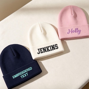 Puede incluir: Tres gorros de punto con dobladillo. Uno es azul marino con texto bordado en azul claro que dice EMBROIDERED TEXT. Uno es crema con texto bordado en negro que dice JENKINS. Uno es rosa claro con texto bordado en morado que dice Holly.
