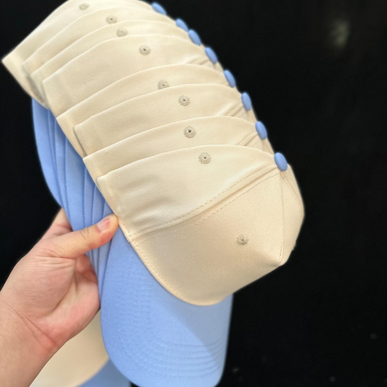 Op de afbeelding: Een stapel baseballpetten met lichtbeige kronen, lichtblauwe knopen aan de bovenkant en lichtblauwe kleppen. Elke petkroon is voorzien van een klein rond ventilatieoogje in het midden. De petten zijn in een verticale opstelling op elkaar gestapeld.