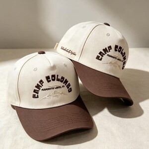 Puede incluir: Dos gorras de b&eacute;isbol con corona color crema y visera marr&oacute;n oscuro. Los paneles frontales presentan un bordado en marr&oacute;n oscuro con el texto CAMP COLOMBO MAMMOTH LAKES, CA sobre un gr&aacute;fico de monta&ntilde;as. Cada gorra tiene un bot&oacute;n marr&oacute;n arriba.