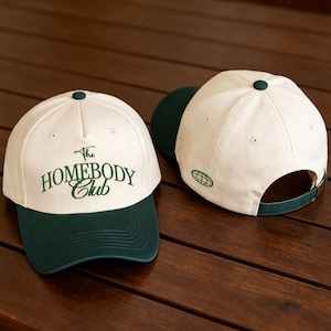 May include: Two baseball caps with cream-colored crowns and dark green brims. The front cap features the dark green embroidered text The HOMEBODY Club. The back cap shows a small green globe icon on the side and an adjustable green strap.