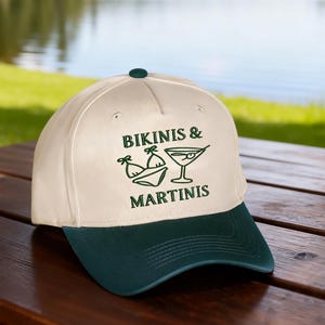 Puede incluir: Una gorra de b&eacute;isbol de color crema con visera verde oscuro y un bot&oacute;n superior. La parte delantera presenta un bordado en verde oscuro de un bikini y una copa de martini, con el texto BIKINIS & MARTINIS bordado en una fuente serif verde oscuro.