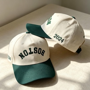 May include: Two baseball caps featuring an off-white crown and a dark green brim. The front of the caps displays the word BOSTON in dark green embroidered lettering. The side of the caps features the embroidered year 2024 in dark green thread.