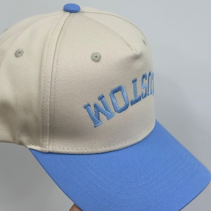 Op de afbeelding: Een beige honkbalpet met een lichtblauwe klep en een bijpassende blauwe knoop aan de bovenkant. Op de voorkant van de pet staat het woord CUSTOM geborduurd in lichtblauw garen. De pet heeft gestikte oogjes en een gestructureerd kroonontwerp.