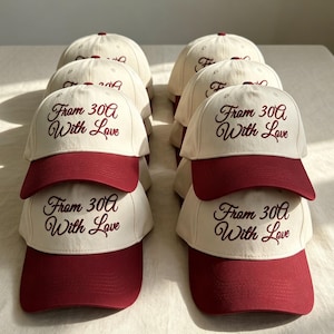 May include: A collection of baseball caps arranged in two rows. Each cap features an off-white crown and a dark red brim. The text 'From 30A With Love' is embroidered in dark red cursive script on the front panel of every cap in the display.