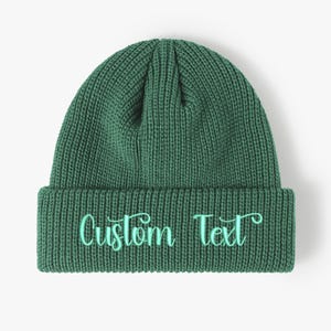Può includere: Un berretto lavorato a maglia verde scuro con bordo ripiegato. Le parole "Custom Text" sono ricamate in uno script verde chiaro sulla parte anteriore del bordo. Il cappello è realizzato in materiale a maglia a coste.