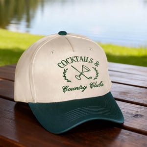 Puede incluir: Una gorra de b&eacute;isbol color crema con visera verde oscuro y un dise&ntilde;o bordado en verde oscuro en la parte delantera. El dise&ntilde;o presenta una copa de martini cruzada con un palo de golf, rodeada de hojas de laurel, con el texto COCKTAILS & Country Clubs.