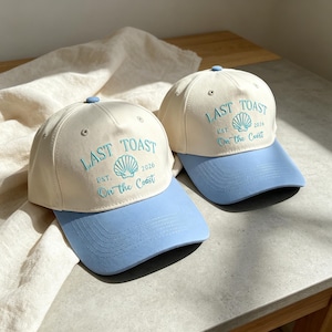 May include: Two baseball caps with cream-colored crowns and light blue brims. Each cap features embroidered teal text that reads LAST TOAST EST. 2026 On the Coast, centered above a small seashell graphic. The caps have a matching light blue button.