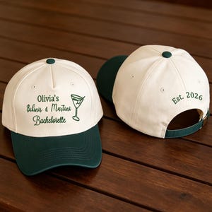 May include: Two baseball caps with cream-colored crowns and dark green brims. The front cap features dark green text reading Olivia's Bikinis & Martini Bachelorette with a martini glass icon. The back cap displays the dark green text Est. 2026.