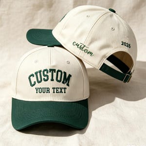 Puede incluir: Dos gorras de béisbol con corona color blanco hueso y visera verde oscuro. La gorra frontal muestra el texto bordado CUSTOM YOUR TEXT en verde oscuro. La gorra trasera presenta la palabra custom y el número 2026 bordados en verde oscuro.