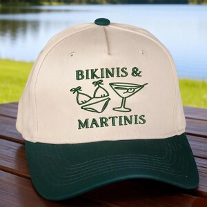 Puede incluir: Una gorra de b&eacute;isbol de color crema con visera verde oscuro y un bot&oacute;n verde oscuro en la parte superior. El frente presenta un bordado verde oscuro de un bikini y una copa de martini, con el texto BIKINIS & MARTINIS bordado en letras verdes.