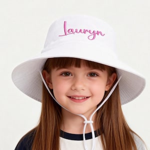 May include: A white bucket hat featuring the name Lauryn embroidered in pink script lettering on the front. The hat includes a wide brim and a white adjustable chin strap with a sliding toggle to secure the fit under the chin.