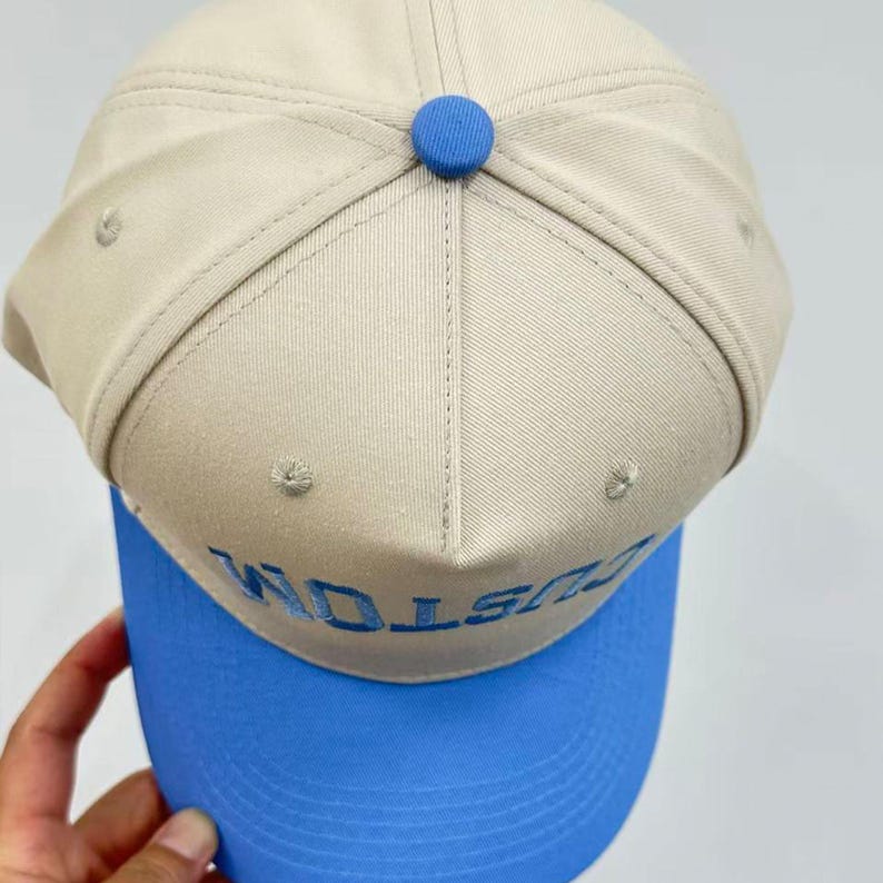 Op de afbeelding: Een beige honkbalpet met een felblauwe klep en een bijpassende blauwe knoop bovenop. Op het voorpaneel staat het woord CUSTOM geborduurd in blauwe draad. De pet is opgebouwd uit zes panelen en is voorzien van kleine ventilatiegaatjes.
