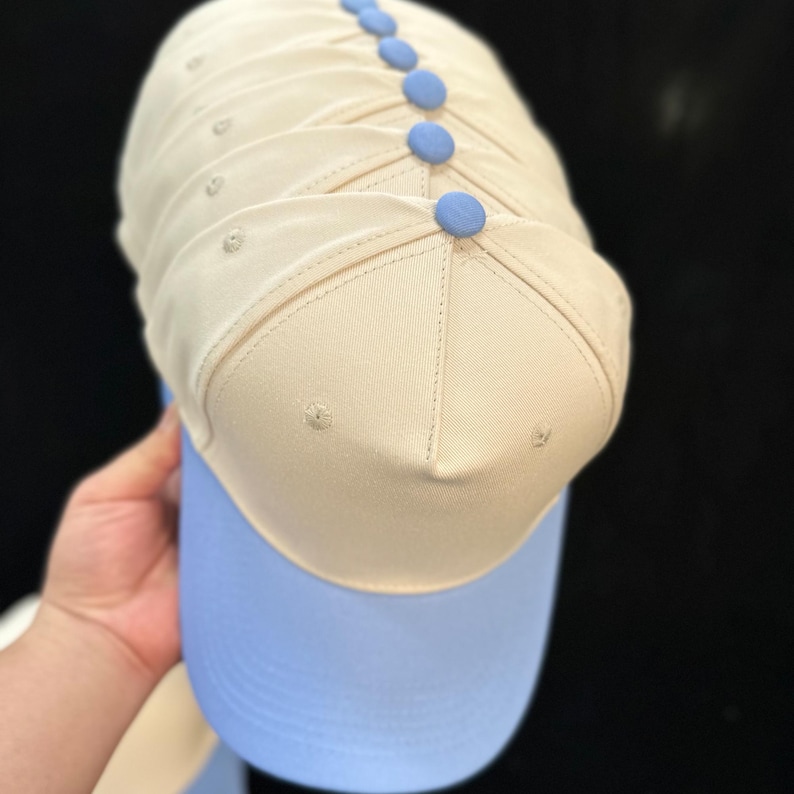 Puede incluir: Una gorra de b&eacute;isbol con una corona de color beige y una visera de color azul claro. La corona est&aacute; construida con varios paneles con ojales cosidos y est&aacute; rematada con un peque&ntilde;o bot&oacute;n redondo de color azul claro en el v&eacute;rtice superior.