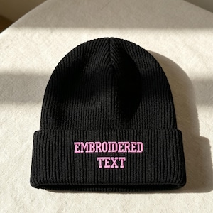 May include: A black ribbed knit beanie featuring a folded cuff. The front of the cuff displays the words EMBROIDERED TEXT stitched in a light pink thread using a bold, block-style font. The hat is constructed from a thick, textured yarn material.