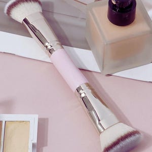 Könnte beinhalten: Ein doppelseitiger Make-up-Pinsel mit rosa Griff und silbernen Akzenten. Die Pinselköpfe sind weiß mit braunen Spitzen. Im Hintergrund befindet sich eine quadratische Foundation-Flasche mit schwarzem Deckel sowie eine kleine Lidschattenpalette.