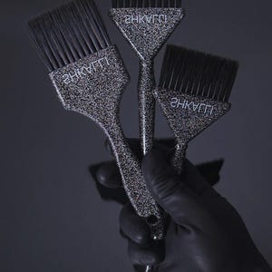May include: Three professional hair dye brushes with black bristles and glittery silver handles. Each brush has the word "2HKVTT" printed on it in white. The brushes are held by a gloved hand against a dark background.