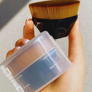 Könnte beinhalten: Ein schwarzer und brauner Make-up-Pinsel mit einer transparenten Kunststoffhülle. Der Pinsel hat einen flachen, abgewinkelten Kopf mit weichen, hellbraunen Borsten. Die Hülle ist rechteckig und hat einen Klappdeckel. Der Pinsel wird in einer Hand gehalten.