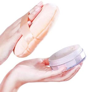 May include: A peach-coloured powder puff with a satin ribbon handle is held above a clear, round container of loose powder. The container has a light pink lid. The background is white.