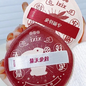 May include: Two peach-shaped cosmetic puffs. One is red, the other is pink. Both have the text "ixix" and Chinese characters. Each puff has a white ribbon with Chinese text. The red puff has the text "量天求好".