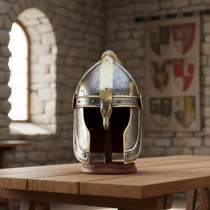 May include: A detailed, antique-style helmet with a silver and gold finish, displayed on a wooden stand. The helmet features intricate designs and a decorative gold accent on the front. The background includes a wooden table and a stone wall.