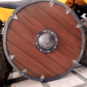 May include: A round wooden shield with a dark brown finish and a metal rim. The shield features a central boss and decorative metal accents. The shield's diameter is approximately 30 inches.