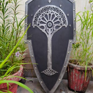 May include: A black shield with a silver tree design, framed with silver and rivets. The tree has a detailed, stylized design. The shield is displayed outdoors with plants in the background.