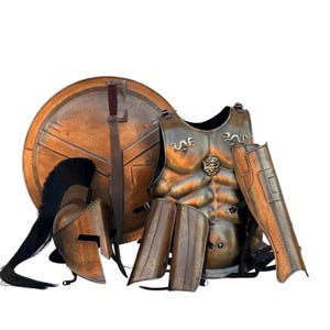 May include: A bronze-toned metal armor set including a circular shield, a Corinthian-style helmet with a black crest, a chest plate featuring a central lion head emblem, a sword, and matching leg greaves with black leather straps and buckles.