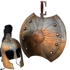 Könnte beinhalten: Ein metallischer, bronzefarbener Helm im Spartaner-Stil mit schwarzem Helmbusch und ein passender großer, runder Schild mit einem zentralen geprägten Medaillon, strahlenförmigen Blütenmustern und kleinen Nieten am gewellten Rand.