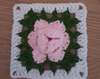 May include: A square, handmade crochet piece featuring a central pink rose design. The rose is surrounded by green leaves, all set against a white square border. The piece is likely a decorative item or part of a larger craft project.
