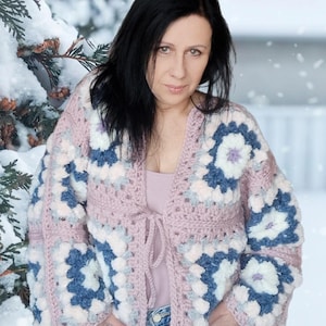 May include: A handmade, open-front cardigan in a granny square design. The sweater features a soft pink base color with large floral squares in shades of blue, white, and lavender. It has long sleeves and a tie closure at the waist, perfect for layering.