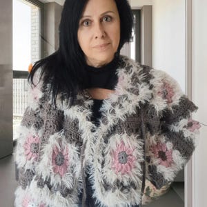 May include: A handmade crochet jacket featuring a floral design in shades of gray, white, and pink. The jacket has a textured, fluffy trim and is worn over a black top and blue jeans. The jacket is a unique piece of wearable art.