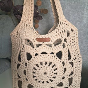 Könnte beinhalten: Eine cremefarbene, handgefertigte Häkeltasche mit großem Blumenmuster. Die Tasche hat eine rechteckige Form mit einer weiten Öffnung und zwei Henkeln. Ein kleines Lederetikett mit dem Wort "handmade" ist angebracht.