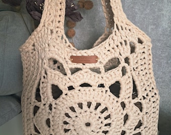 Op de afbeelding: Een crèmekleurige, handgemaakte gehaakte tote bag met een groot bloemenpatroon. De tas heeft een rechthoekige vorm met een brede opening en twee handvatten. Een klein leren label met het woord "handmade" is bevestigd.
